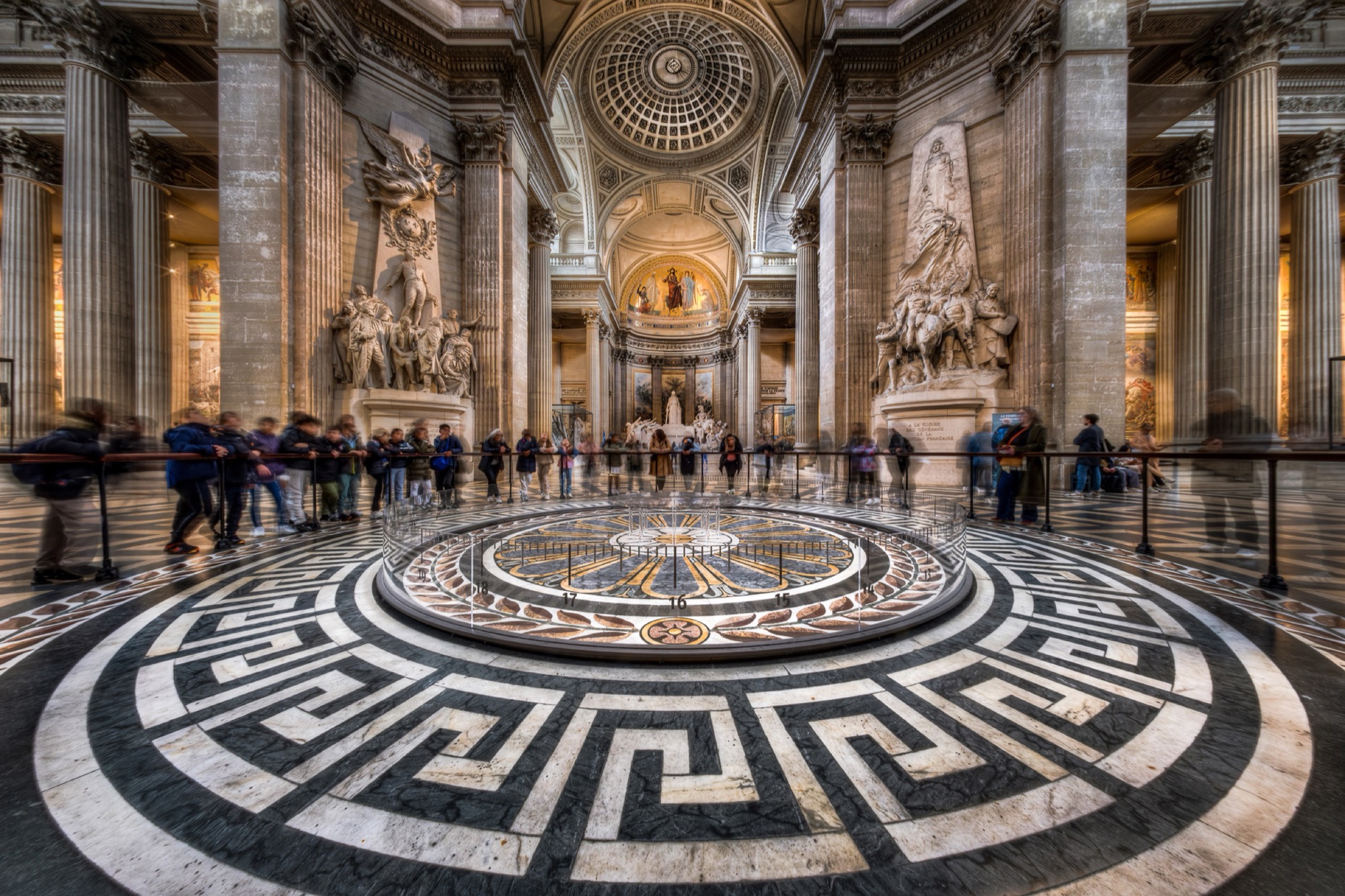 L’intérieur du Panthéon, à Paris : un sol de marbre aux cercles concentriques ornés de grecques, des colonnes et des sculptures à l’arrière-plan, les visiteurs changés en ombres mouvantes.