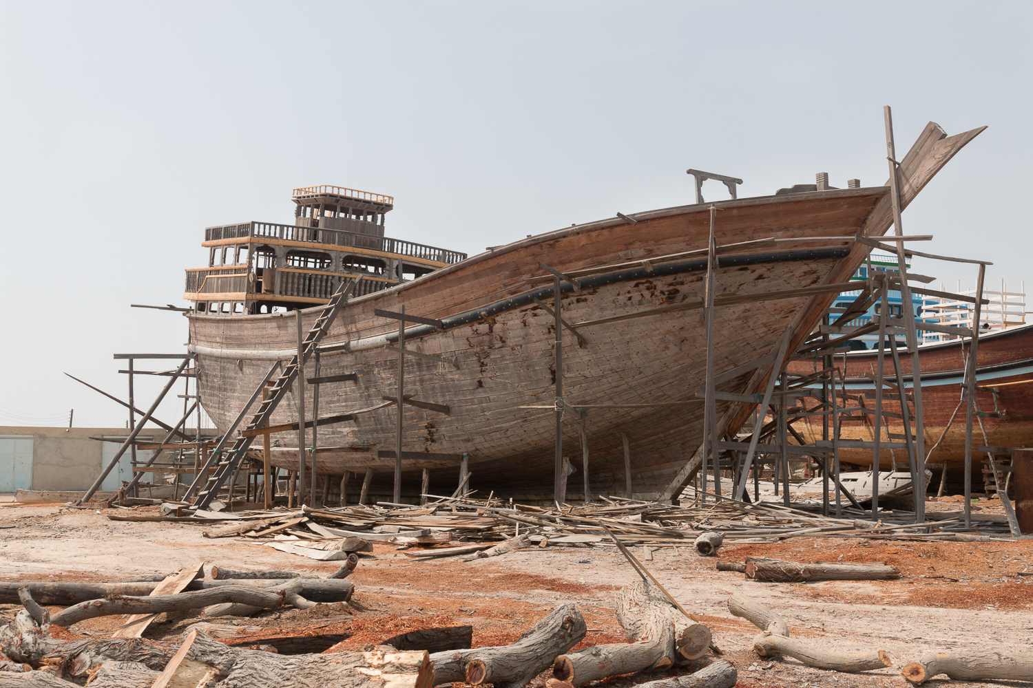 Un grand lenj de bois en construction dans un chantier naval de l’île de Qeshm, soutenu par des échafaudages, des chutes de bois jonchant le sol.