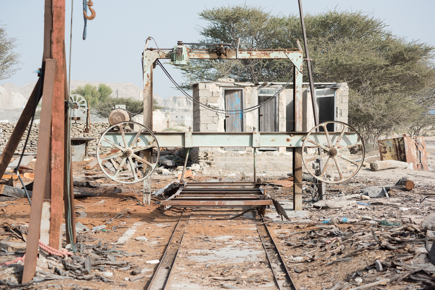 Dans le chantier, une scie à ruban rouillée aux deux grandes roues à rayons enjambe une paire de rails, devant une petite cabane de parpaings et un arbre à la large ramure.