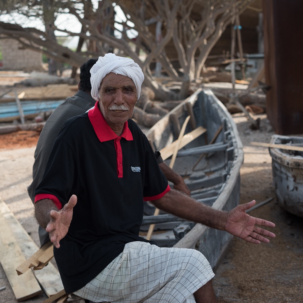 Un homme d’un certain âge, turban blanc et moustache grise, en polo sombre et pagne à carreaux, est assis près d’un petit bateau en bois, dans le chantier, les bras écartés et les paumes ouvertes ; derrière lui, un autre homme est assis, en partie caché.