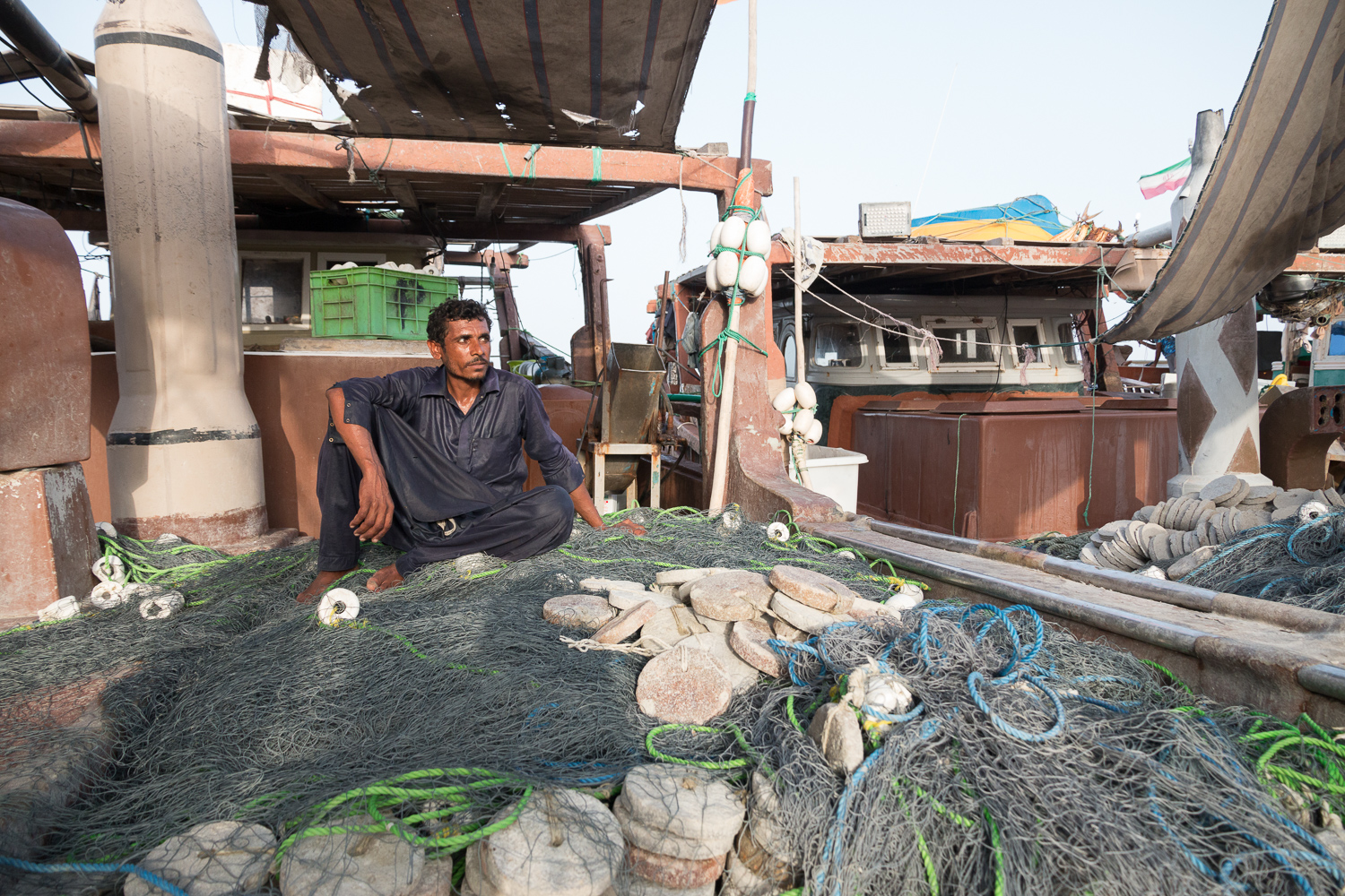 Un homme en tunique bleu foncé est assis pieds nus sur un amas de filets de pêche, de flotteurs blancs et de pierres rondes servant de lest, sur le pont d’un bateau amarré parmi d’autres.