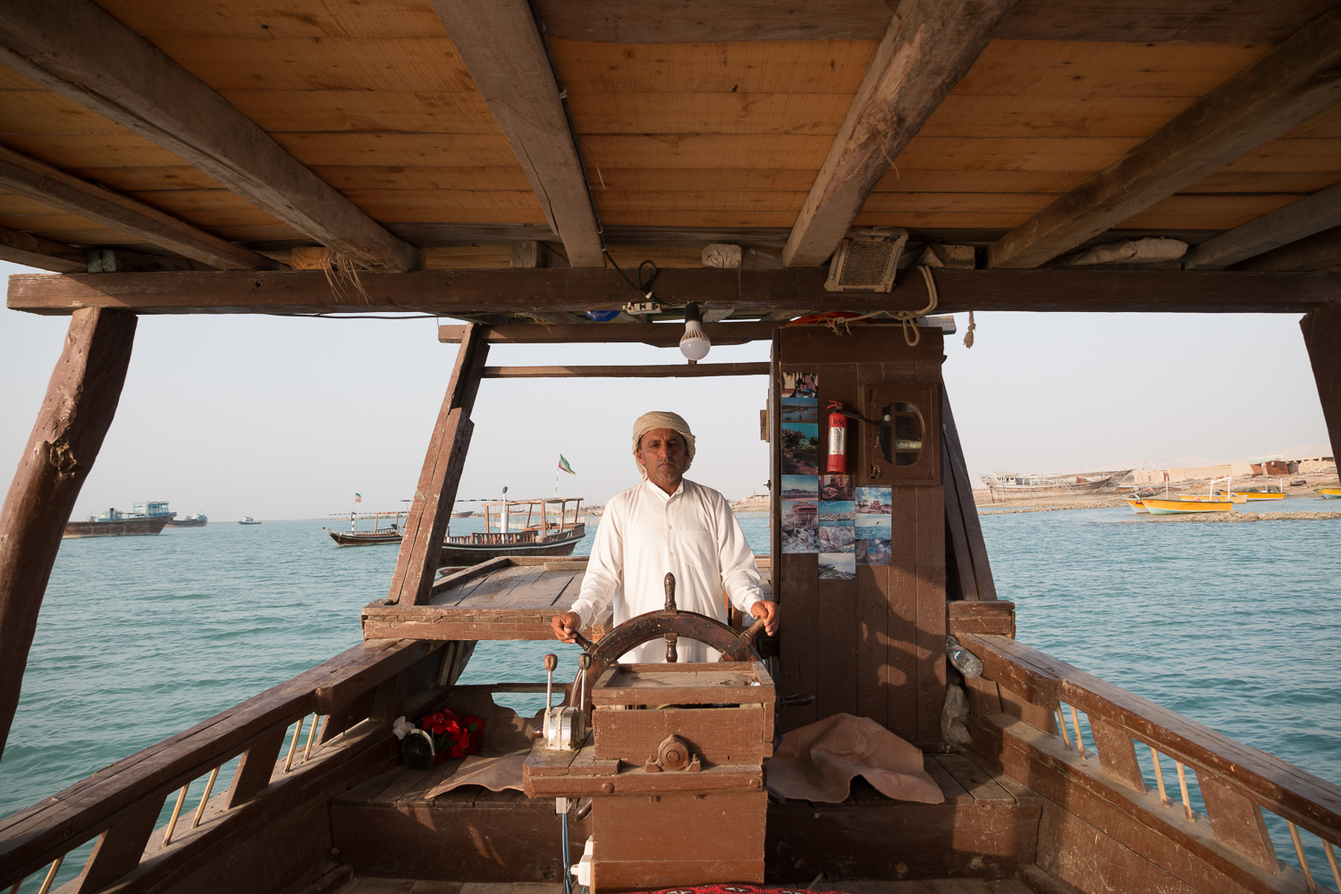 Un homme en robe blanche, la tête couverte d’une étoffe, se tient debout, les deux mains sur la roue à rayons du gouvernail d’un lenj en bois, sous sa toiture de charpente ouverte ; derrière lui, des bateaux mouillent sur une eau calme, non loin du rivage.