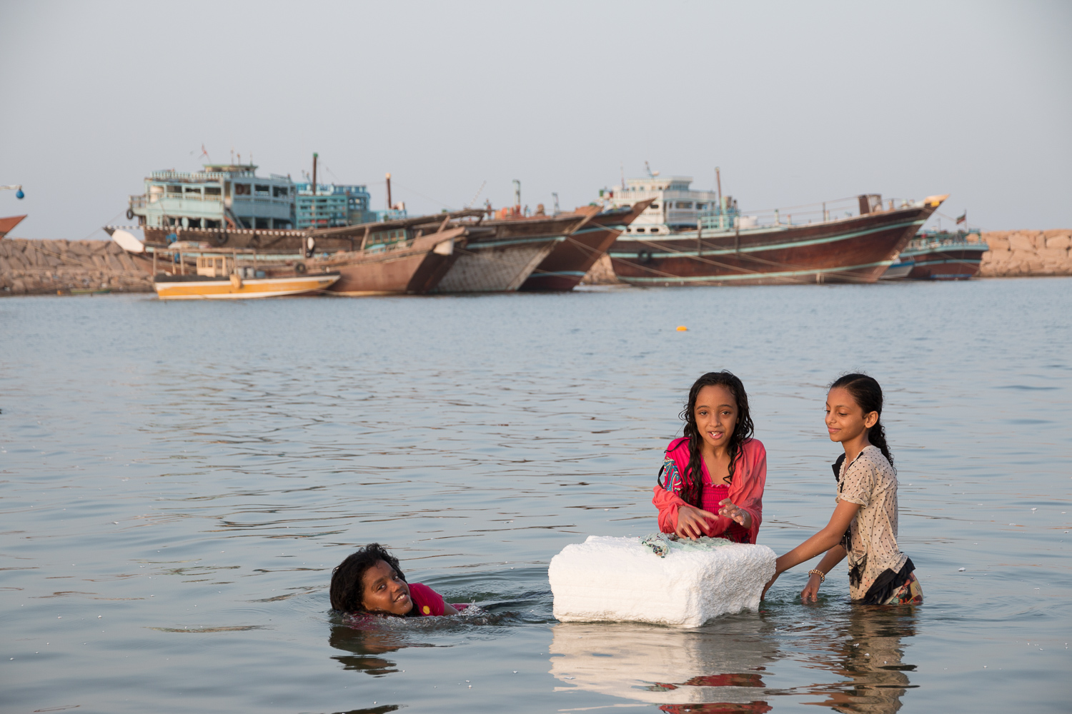 Trois fillettes jouent avec un bloc de polystyrène blanc dans l’eau peu profonde d’un port ; derrière elles, de grands lenjes en bois sont amarrés le long d’une digue de pierre.