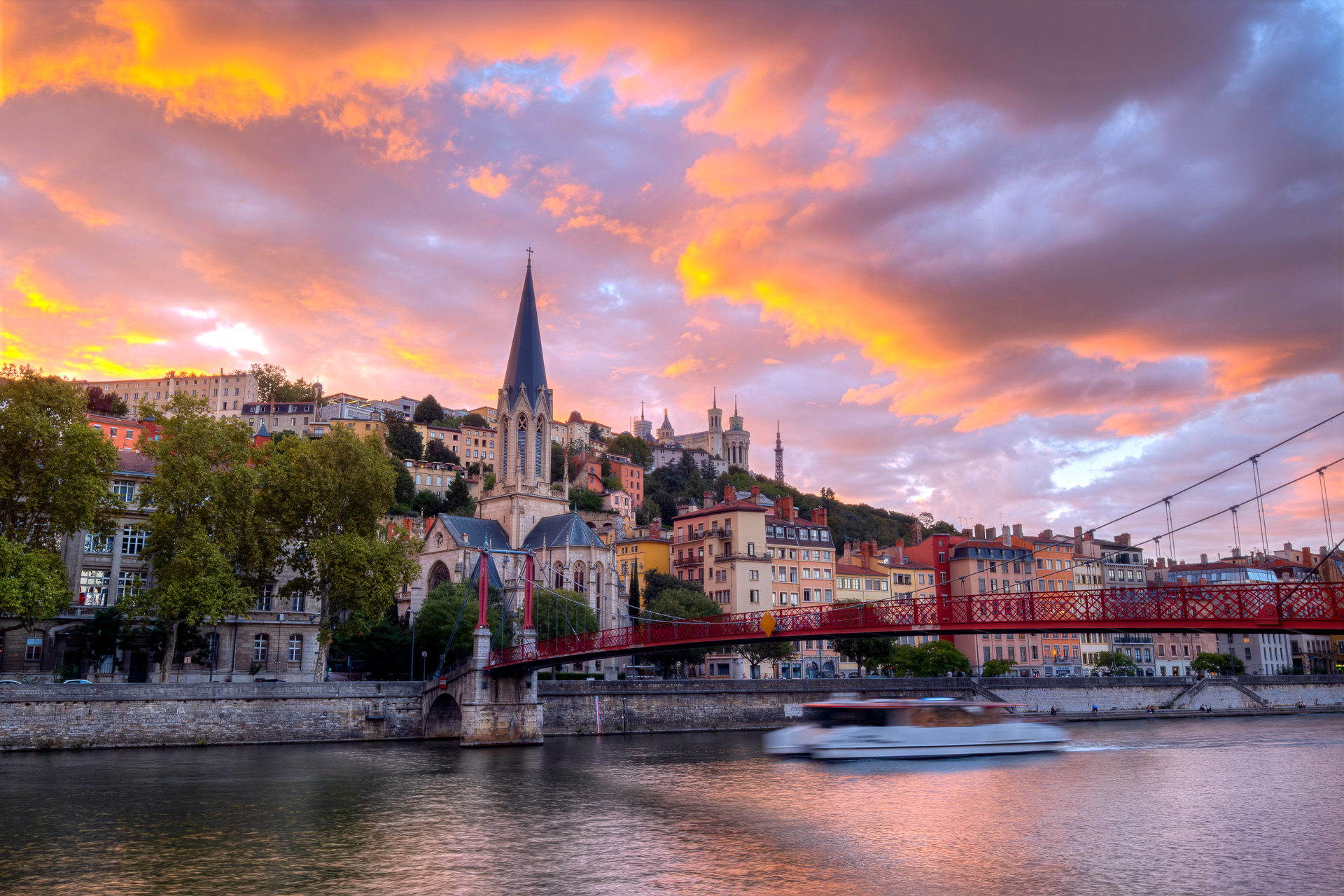 L’église Saint-Georges et la passerelle Saint-Georges rouge sur la Saône sous un ciel de couchant embrasé, la basilique de Fourvière et la tour métallique sur la colline, un bateau passant en filé