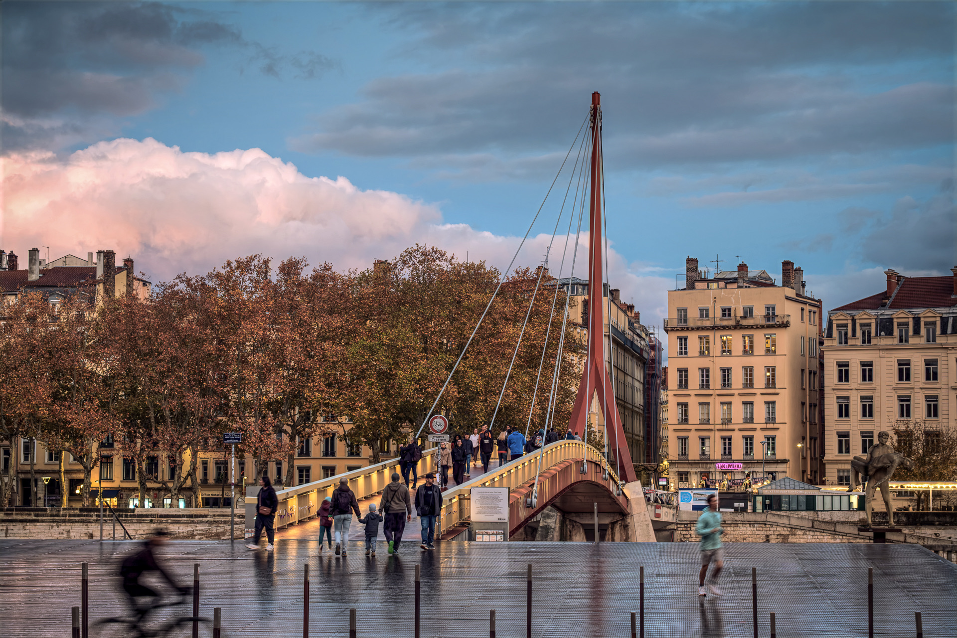 Le mât rouge de la passerelle du Palais de Justice sur la Saône après la pluie, des familles s’engageant sur la passerelle depuis l’esplanade mouillée, un cycliste et un coureur de passage, les platanes d’automne et les quais de la Presqu’île au loin