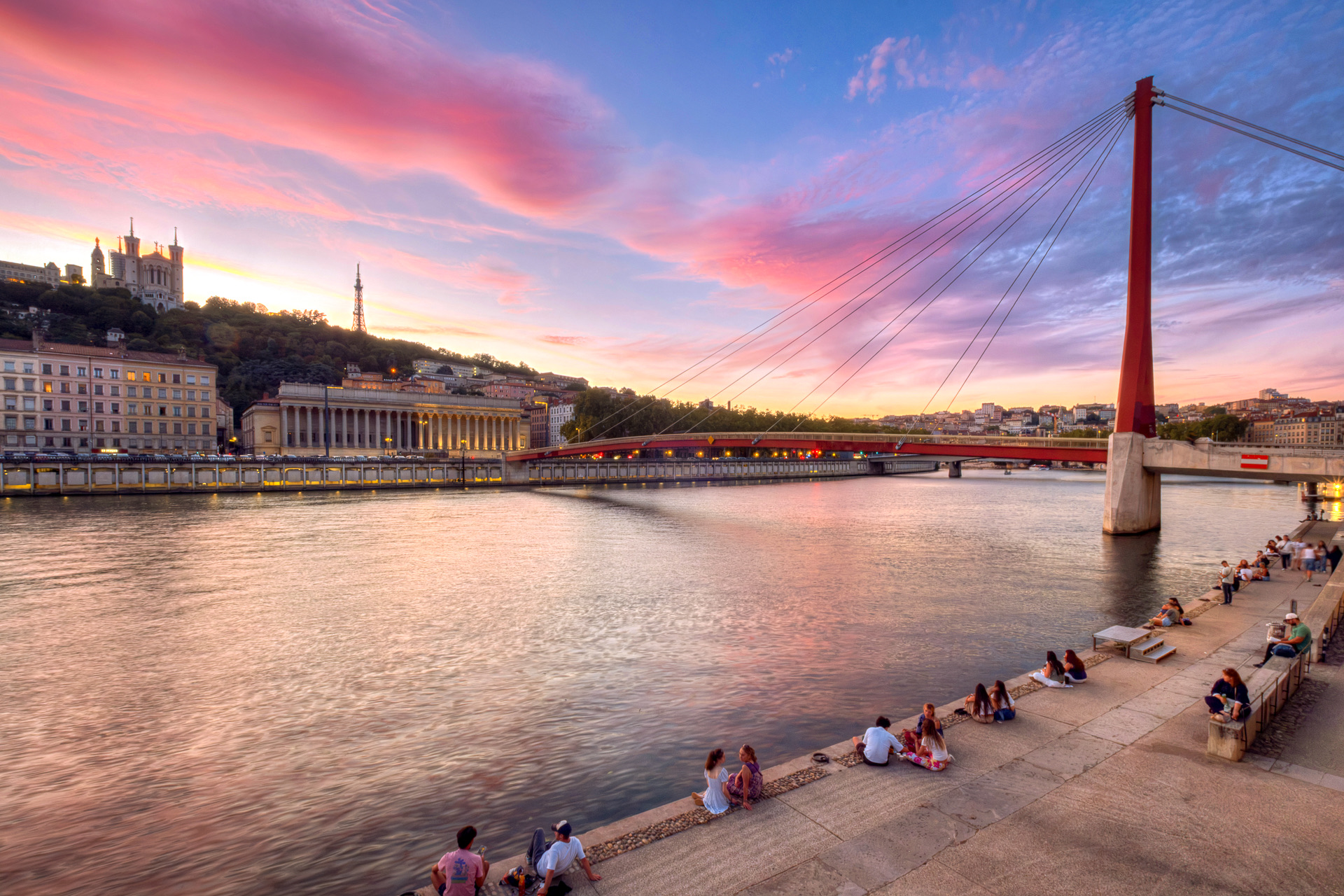 Coucher de soleil sur la Saône : la passerelle rouge du Palais de Justice, le palais des 24 colonnes et Fourvière, des promeneurs assis sur le quai des Célestins
