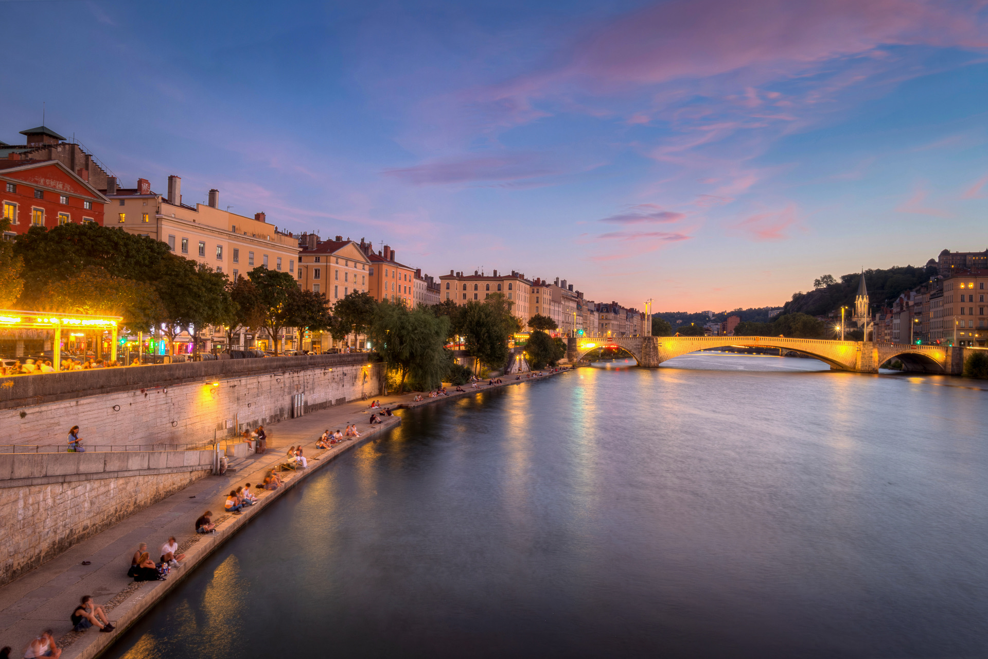 La Saône à l’heure bleue depuis la passerelle du Palais de Justice, vers l’aval : des promeneurs assis sur le bas-port du quai des Célestins, le pont Bonaparte illuminé au loin et la flèche de Saint-Georges sur l’autre rive