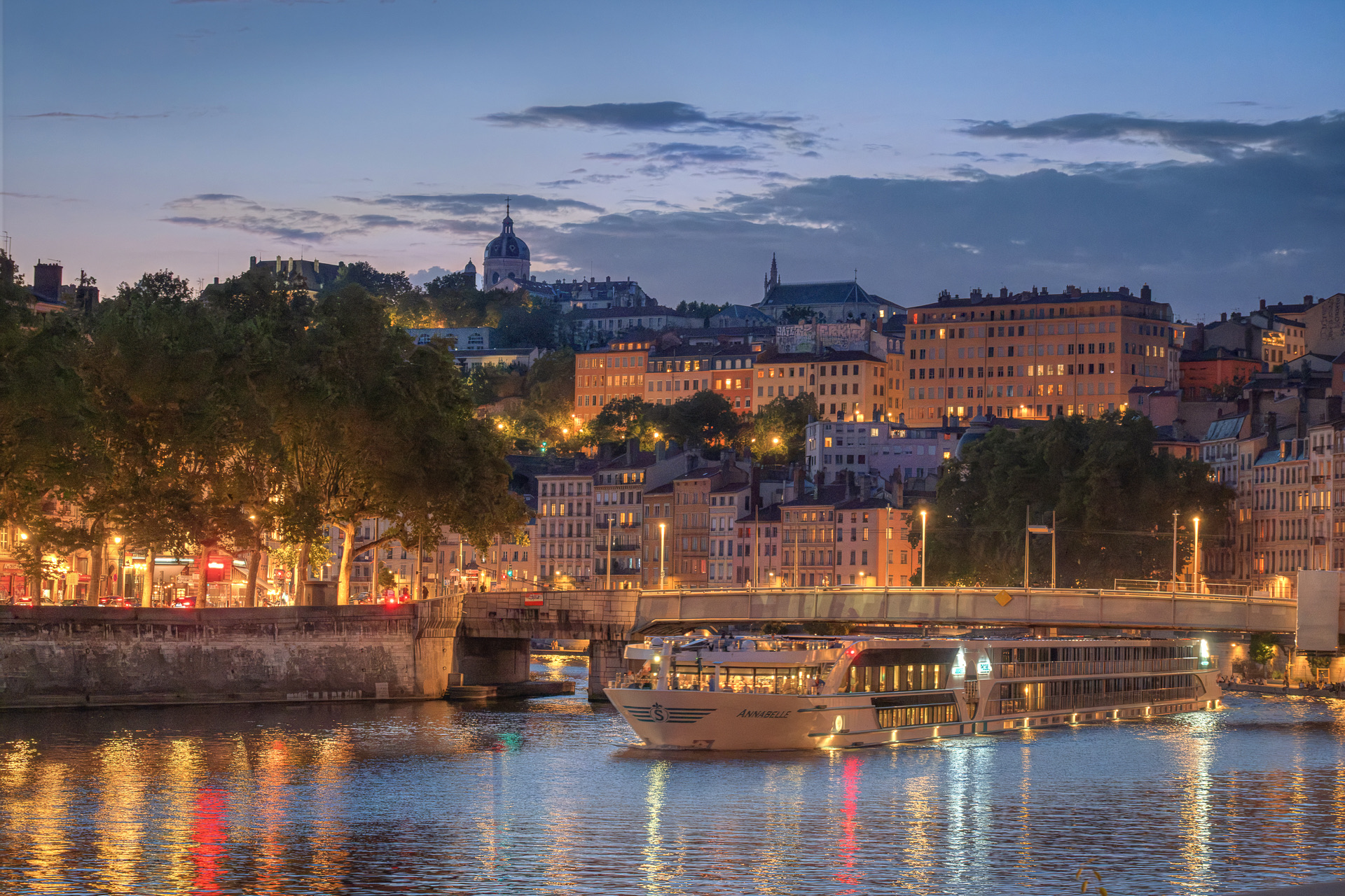 La Saône à l’heure bleue : un bateau de croisière illuminé près du pont La Feuillée, les lumières du quai de Bondy et les maisons éclairées de la pente des Chartreux jusqu’au dôme de Saint-Bruno-les-Chartreux