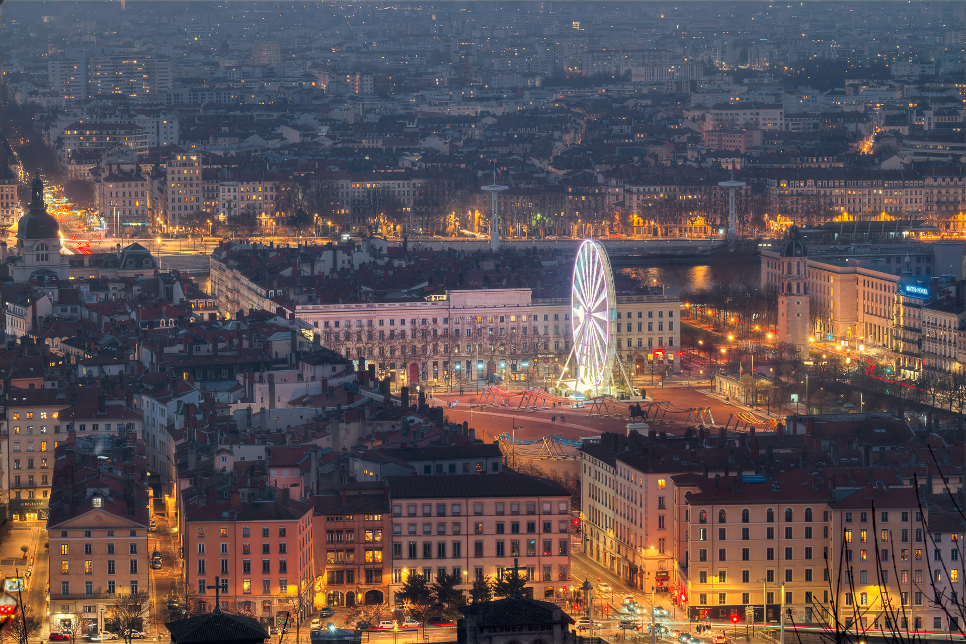 Vue nocturne depuis l’esplanade de Fourvière sur la Presqu’île : la place Bellecour avec sa grande roue illuminée et la statue de Louis XIV, le clocher de la Charité, le Rhône et la ville au-delà, le sommet des tours de Saint-Jean au premier plan