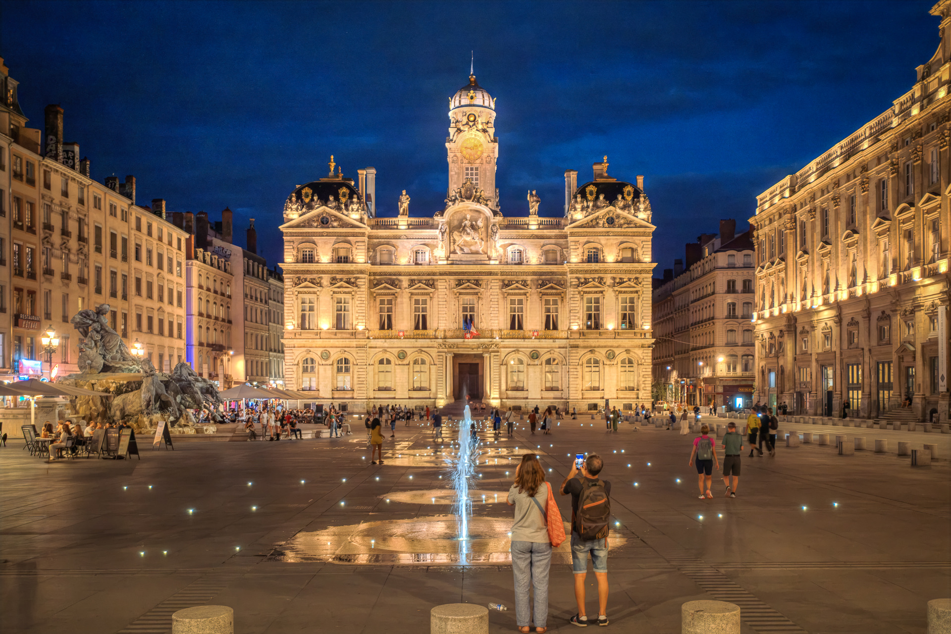 La façade ouest illuminée de l’Hôtel de Ville à l’heure bleue, vue à travers la place des Terreaux : un jet d’eau jaillissant du dallage, la fontaine Bartholdi à gauche, le palais Saint-Pierre à droite, un couple photographiant la scène