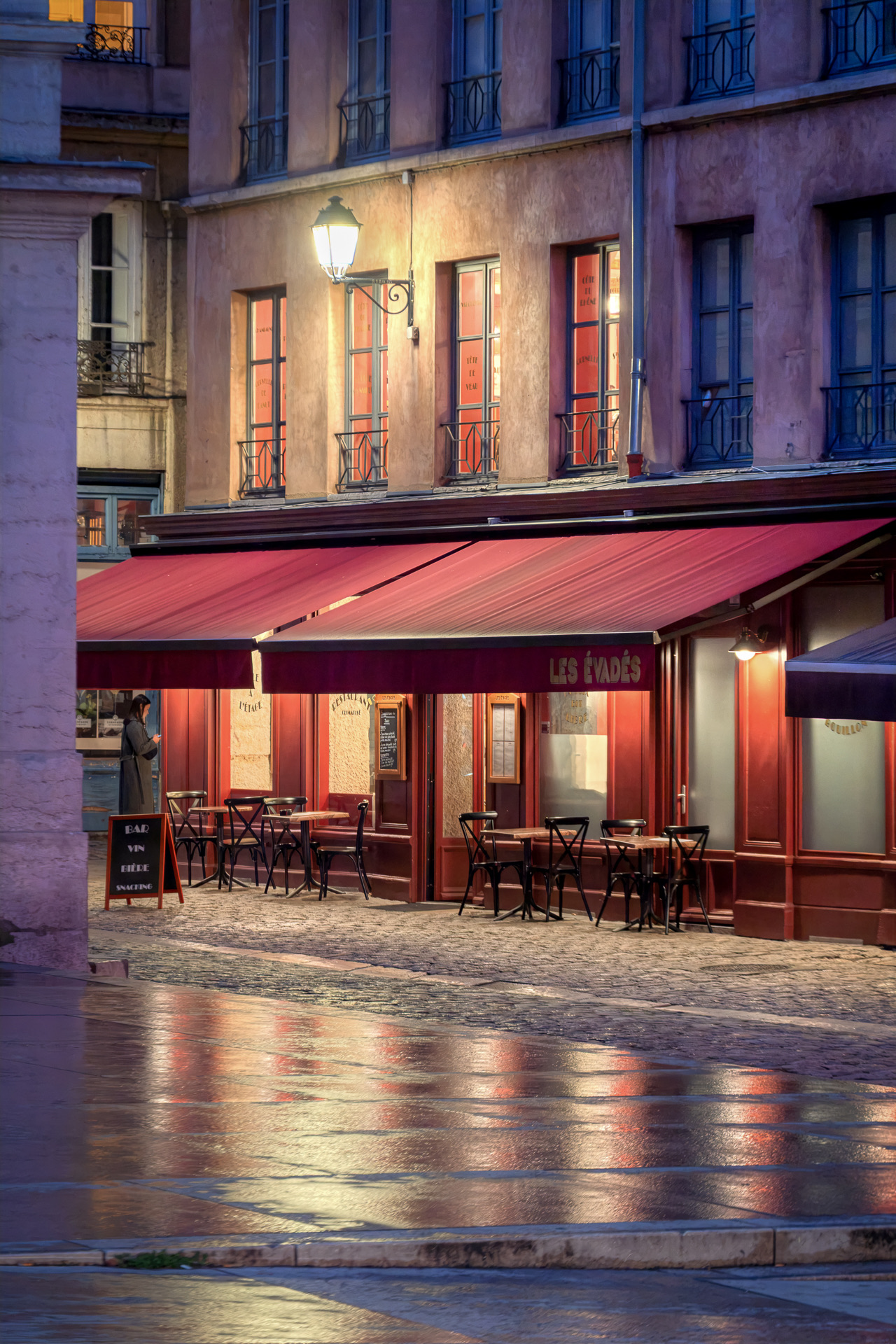 Une brasserie aux stores rouge profond et aux chaises de bistrot vides à l’heure bleue, rue du Palais-de-Justice dans le Vieux Lyon, une lanterne allumée et le pavé mouillé reflétant la lumière rouge