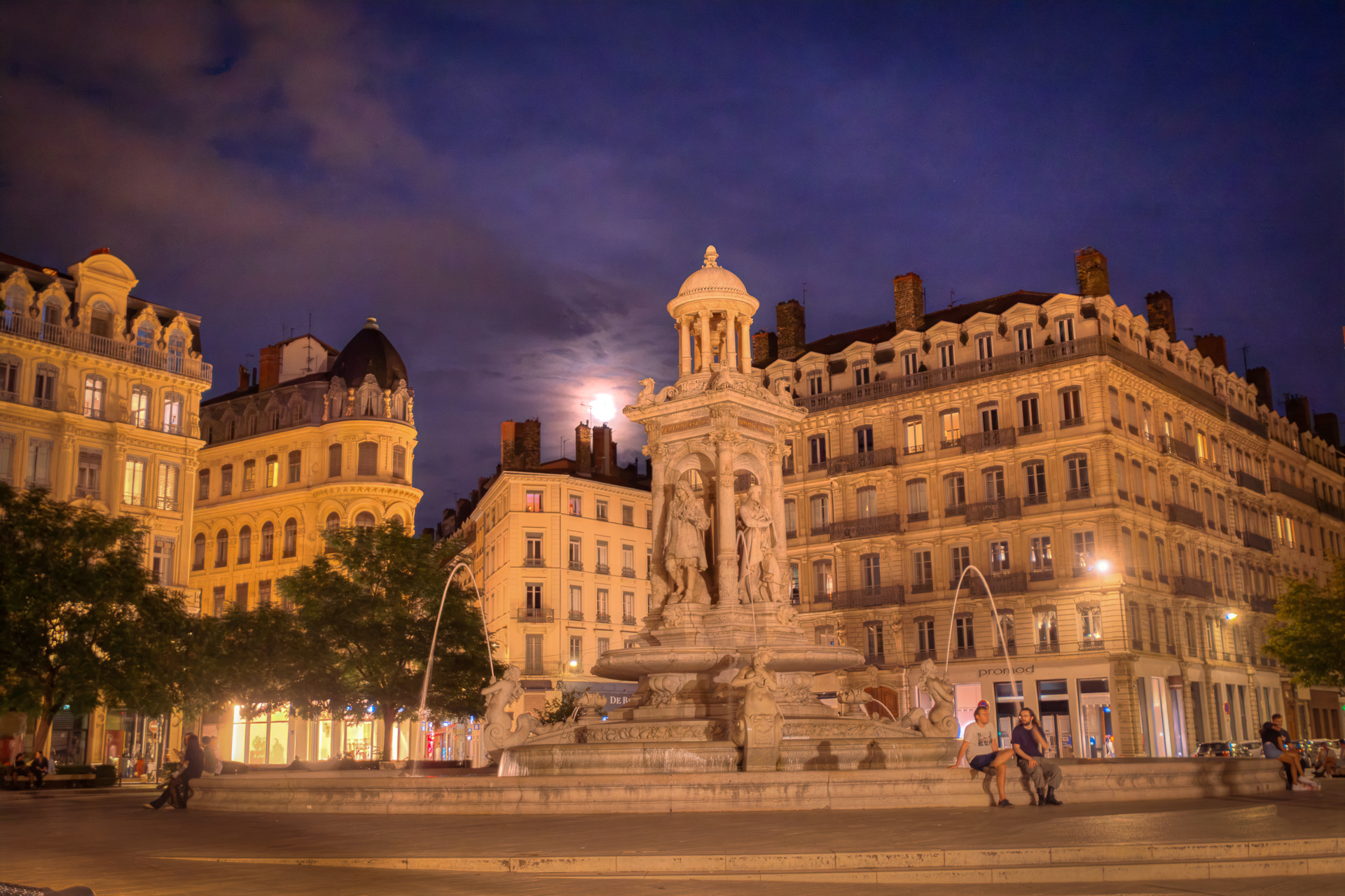 La fontaine des Jacobins en marbre blanc la nuit, ses statues et sa petite rotonde à coupole sur un ciel violet, une lune presque pleine voilée de nuages, les immeubles illuminés de la place des Jacobins