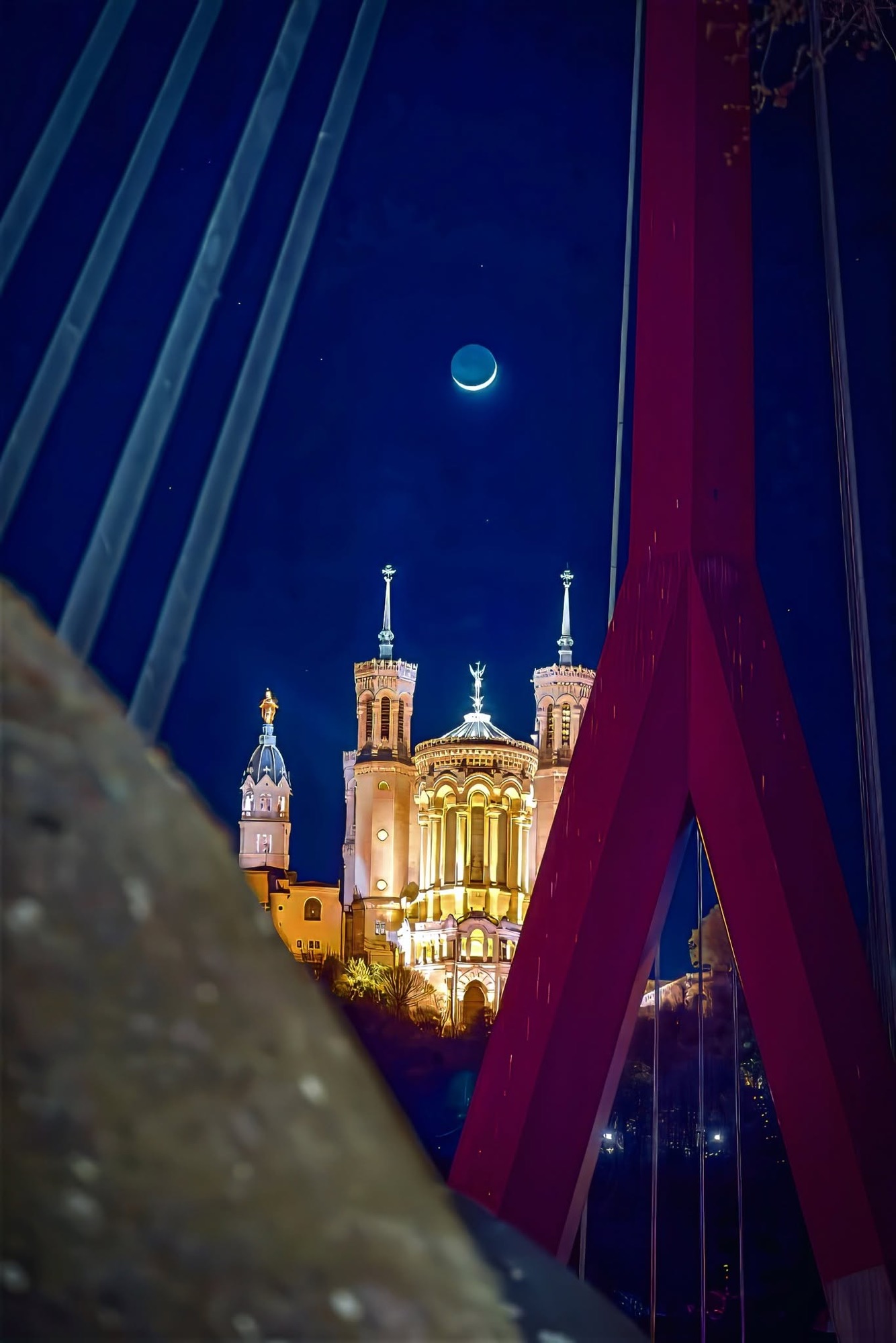Fourvière au clair de lune, encadrée par le pylône rouge et les câbles d’une passerelle