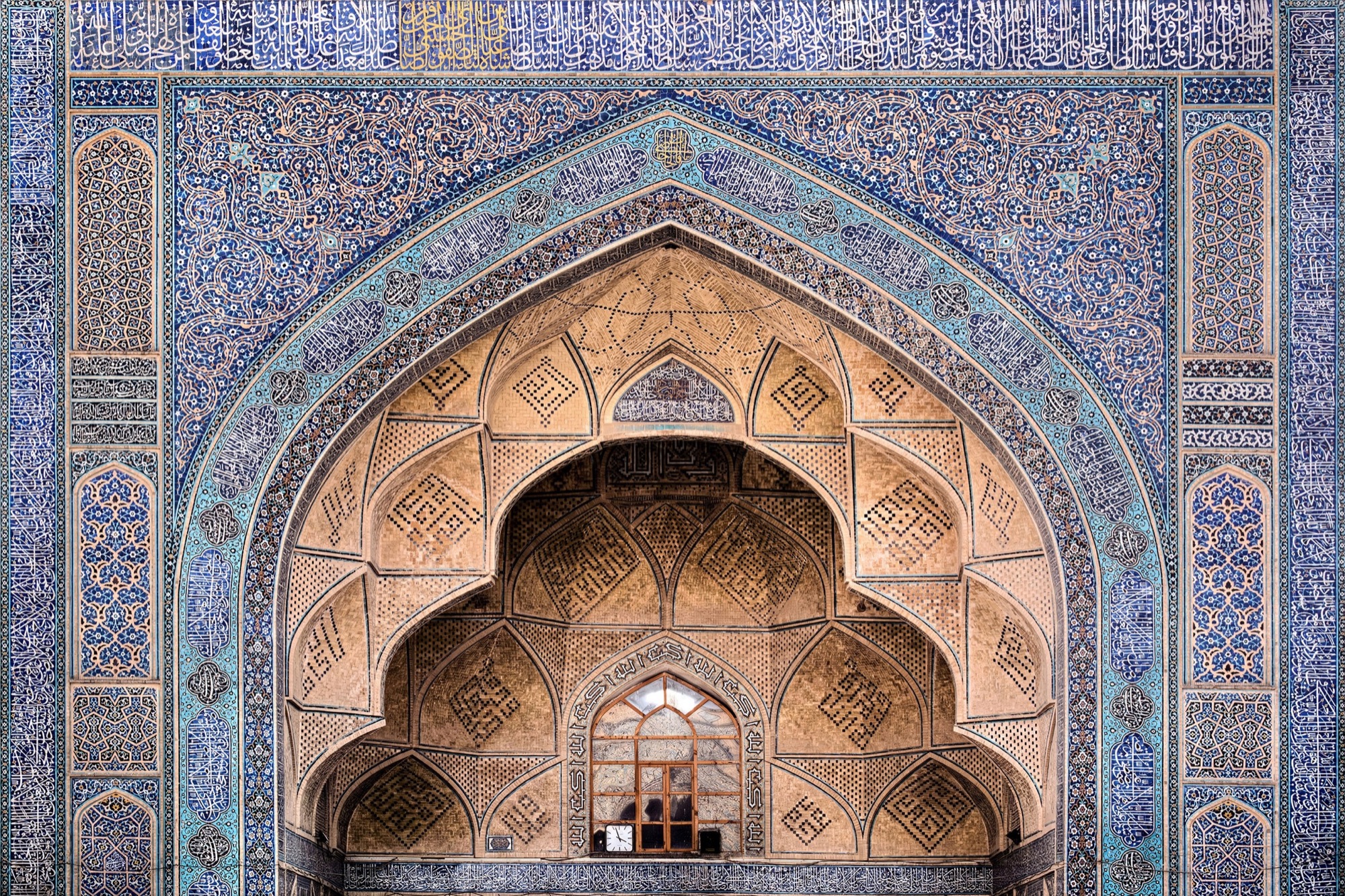 Un portail en arc brisé encadré de céramique bleue, coiffé d’une demi-coupole de muqarnas couleur de miel au-dessus d’une petite fenêtre.