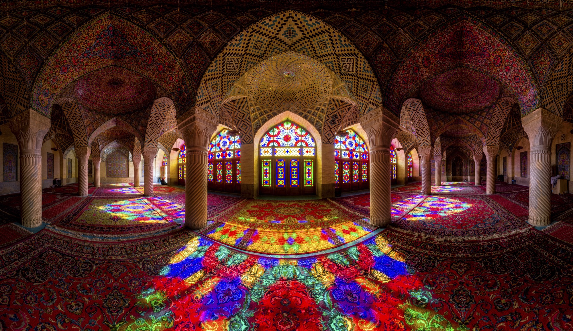 Panorama de la salle de prière de la mosquée Nasir-ol-Molk, à Chiraz : les tapis inondés de la lumière colorée des vitraux, entre des colonnes torsadées.