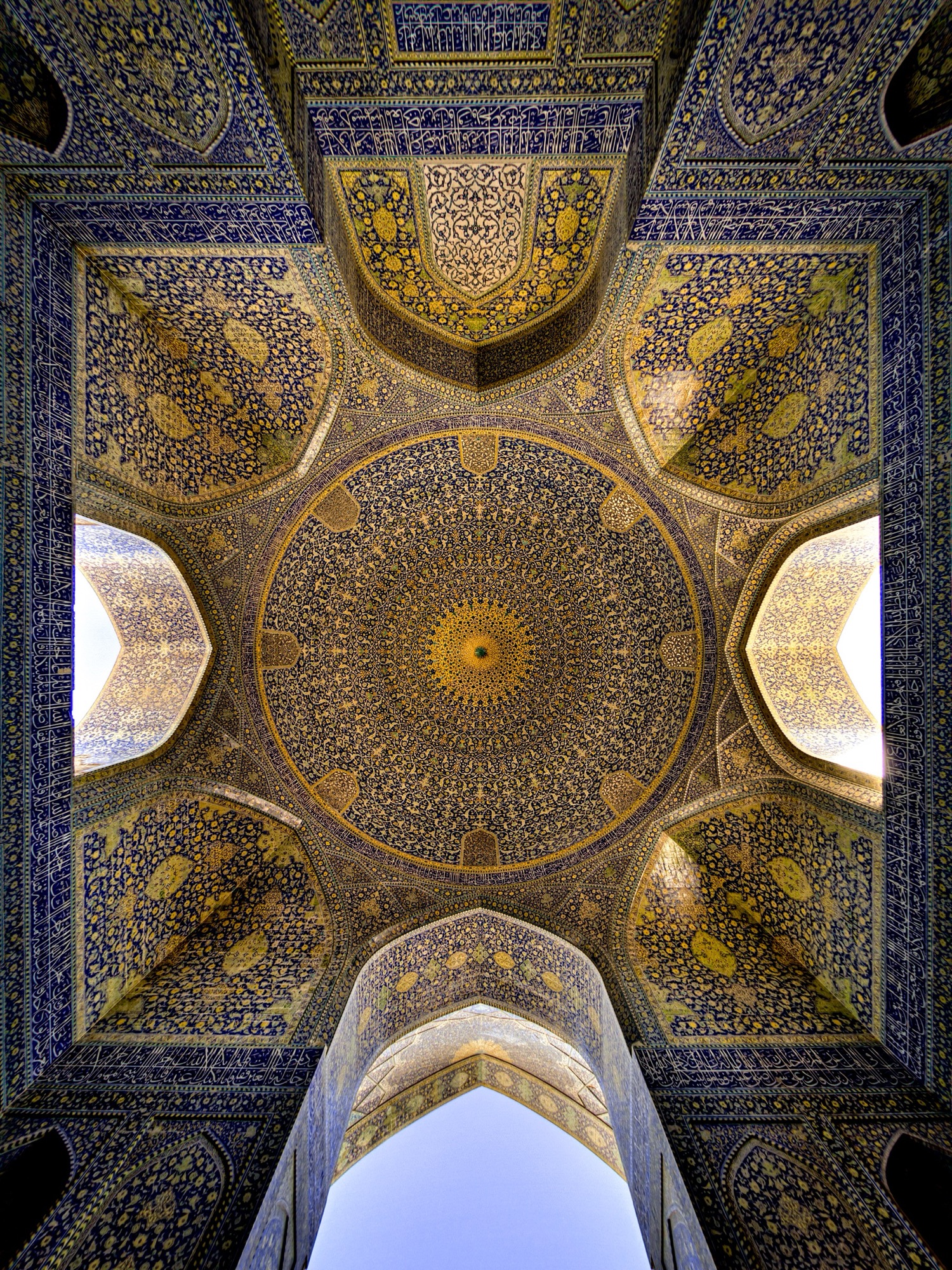 Le plafond d’une salle à coupole de la mosquée Shah Abbasi (mosquée de l’Imam), à Ispahan, vu à la verticale : une coupole de faïence or et bleu au-dessus de quatre angles carrelés et, en contrebas, une arcade ouverte sur le ciel.