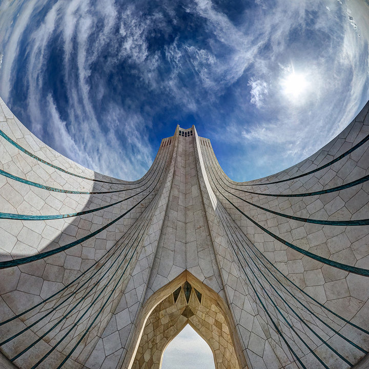 The Azadi Tower in Tehran seen from its foot through a fisheye lens: the white ribbed flanks curve outwards like wings towards a blue sky of swirling cloud and sun.