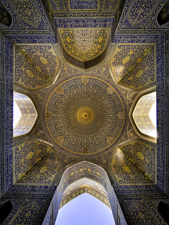 The ceiling of a domed chamber of the Shah (Imam) Mosque in Isfahan, seen straight up: gold and blue tiled vaults around a central dome, with pale sky beyond the arches.