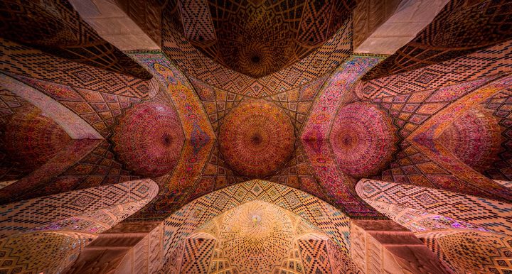 The tiled vaults of the Nasir al-Mulk Mosque in Shiraz, seen from below: rose, red and gold domes and arches in a symmetrical row.