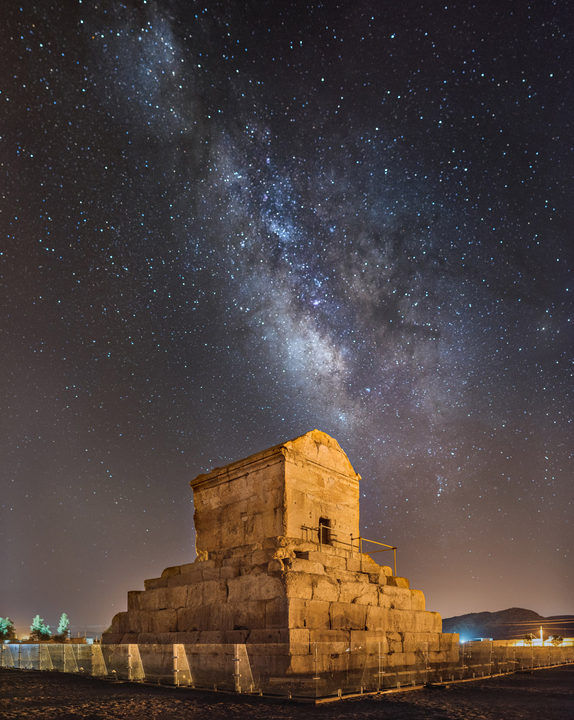 The Tomb of Cyrus the Great at Pasargadae at night: the stone tomb with its gabled chamber on a stepped plinth, lit warm against the dark, beneath the band of the Milky Way.