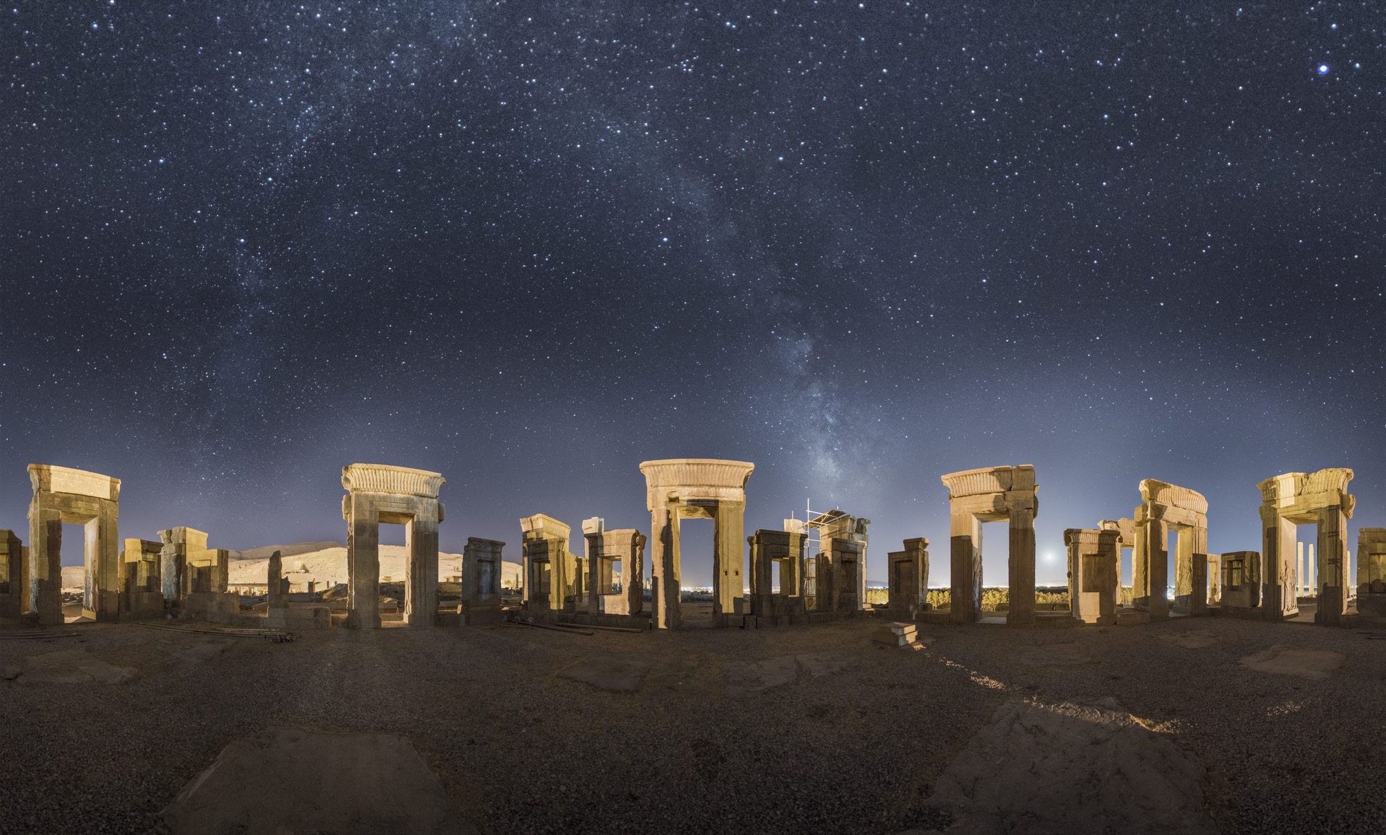 Panorama de Persépolis la nuit : les portes de pierre du palais, éclairées d’une lumière chaude sur un ciel d’un bleu profond, et la Voie lactée qui s’élève à la verticale au-dessus du centre.