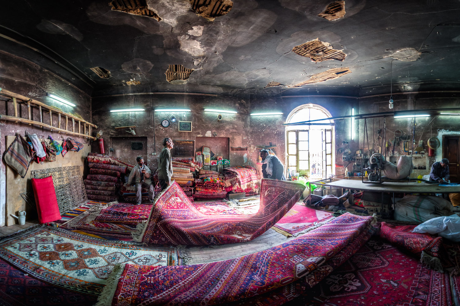 A carpet repair workshop: craftsmen at work among piles of Persian carpets under a high, worn ceiling.